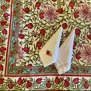 Puede incluir: Un mantel blanco con un patr&oacute;n floral con granadas rojas y hojas verdes. El mantel tiene un borde azul y rojo. Dos servilletas blancas se colocan encima del mantel.