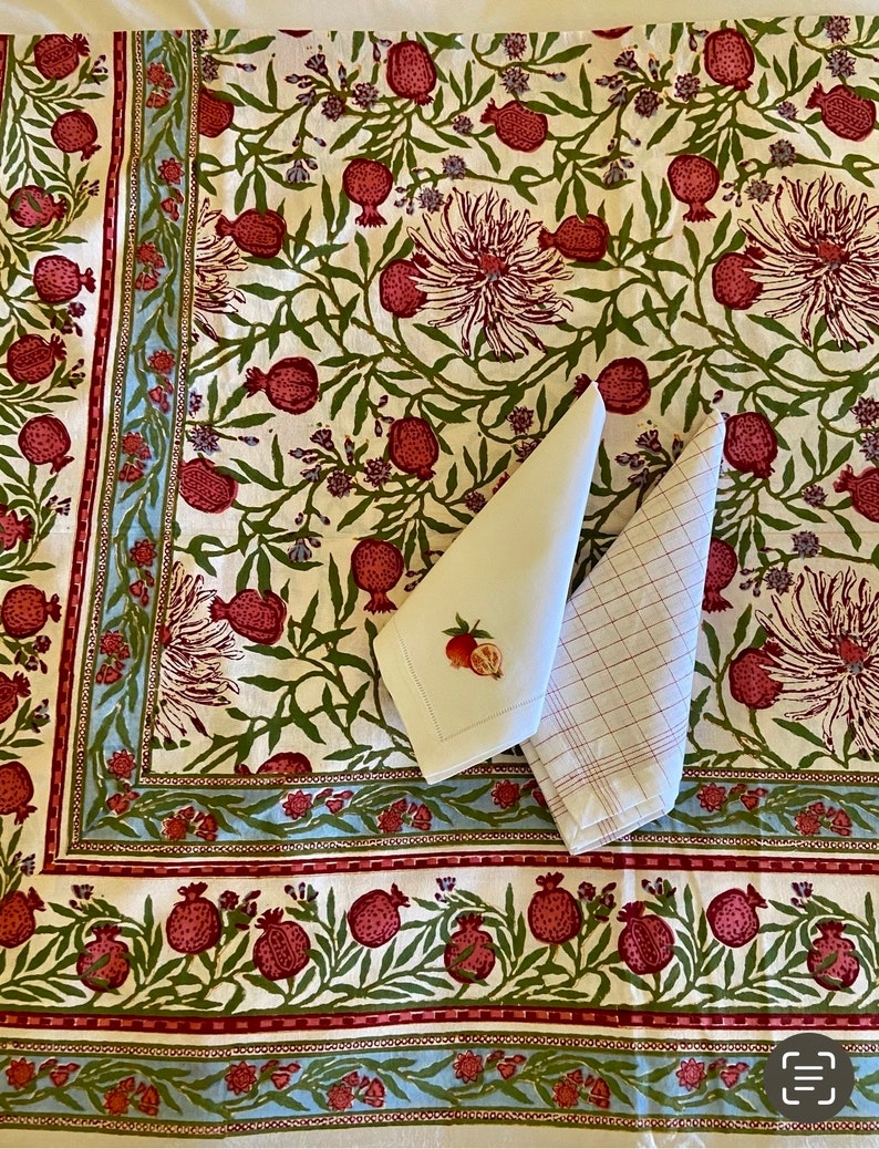 Puede incluir: Un mantel blanco con un patr&oacute;n floral con granadas rojas y hojas verdes. El mantel tiene un borde azul y rojo. Dos servilletas blancas se colocan encima del mantel.