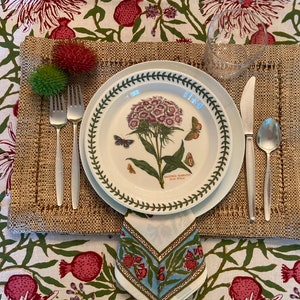 Puede incluir: Un plato blanco con un borde verde que presenta una ilustraci&oacute;n bot&aacute;nica de una flor rosa con hojas verdes y mariposas. El plato est&aacute; colocado sobre un mantel individual tejido marr&oacute;n con una servilleta blanca y cubiertos de plata.