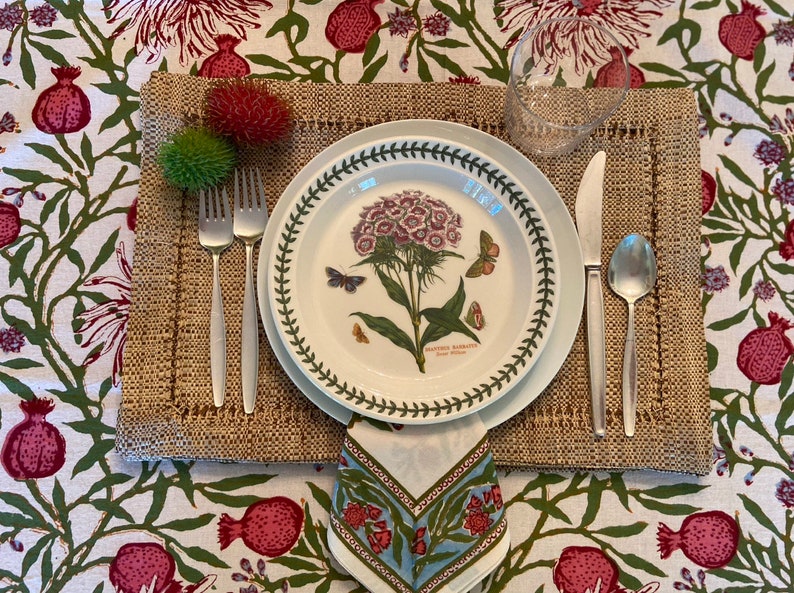 Puede incluir: Un plato blanco con un borde verde que presenta una ilustraci&oacute;n bot&aacute;nica de una flor rosa con hojas verdes y mariposas. El plato est&aacute; colocado sobre un mantel individual tejido marr&oacute;n con una servilleta blanca y cubiertos de plata.