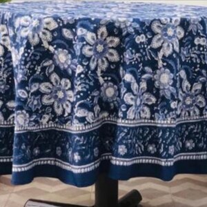 Peut inclure: Nappe ronde avec un motif floral bleu fonc&eacute;. Le motif pr&eacute;sente des fleurs et des feuilles blanches et bleu clair. La nappe est drap&eacute;e sur une table, avec une bordure bleu fonc&eacute; en bas. La table est entour&eacute;e de plantes en pot.