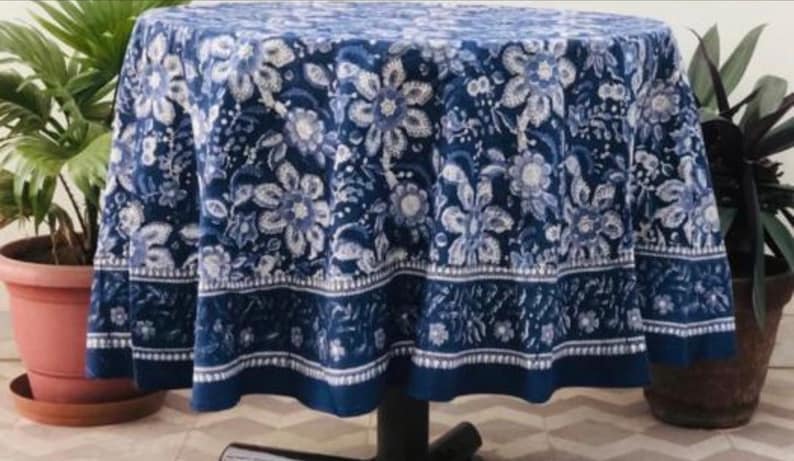 Peut inclure: Nappe ronde avec un motif floral bleu fonc&eacute;. Le motif pr&eacute;sente des fleurs et des feuilles blanches et bleu clair. La nappe est drap&eacute;e sur une table, avec une bordure bleu fonc&eacute; en bas. La table est entour&eacute;e de plantes en pot.