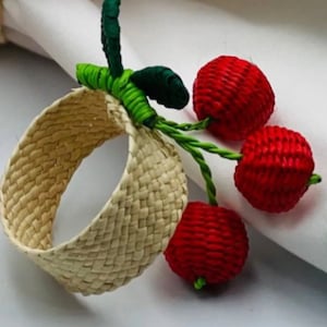 May include: A woven straw napkin ring with three red cherries attached. The cherries are made of woven material and have green stems and leaves.