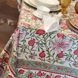 Puede incluir: Mantel blanco con un patr&oacute;n floral con granadas rojas y hojas verdes. El mantel tiene un borde azul y rojo. El mantel est&aacute; puesto con platos, cuencos y tazas blancas.