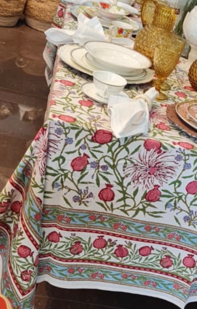 Puede incluir: Mantel blanco con un patr&oacute;n floral con granadas rojas y hojas verdes. El mantel tiene un borde azul y rojo. El mantel est&aacute; puesto con platos, cuencos y tazas blancas.