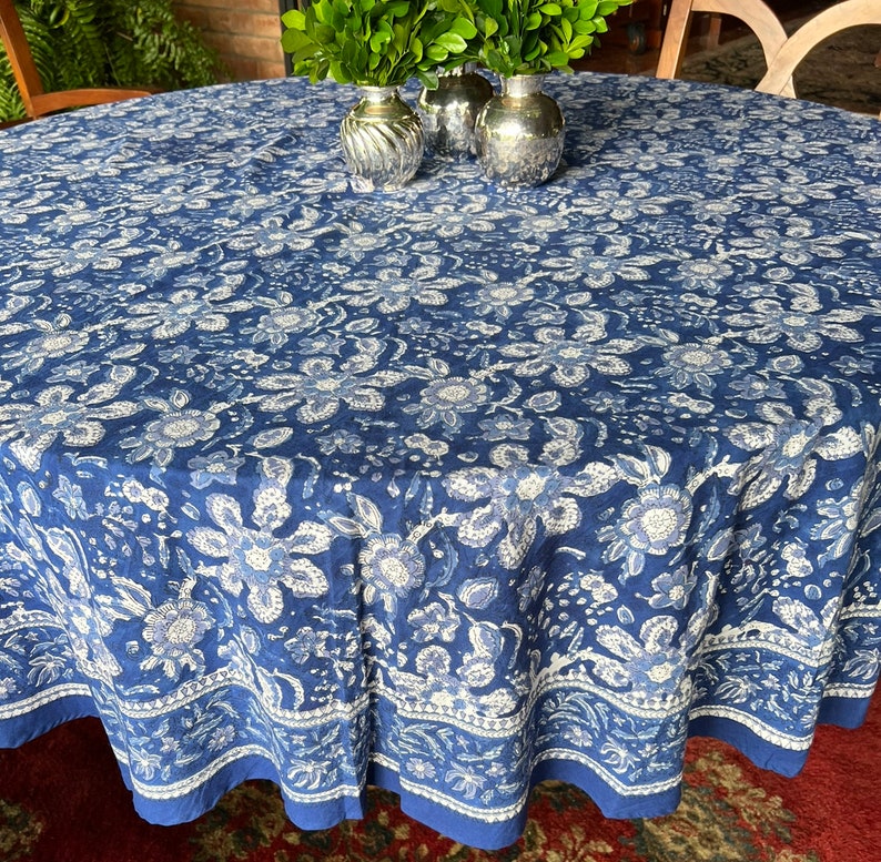 Peut inclure: Une nappe ronde avec un motif floral bleu. La nappe pr&eacute;sente un fond bleu fonc&eacute; avec des motifs floraux blancs et bleu clair. Le bord a une bordure blanche avec des motifs floraux et une bordure bleue unie. La nappe est sur une table ronde.
