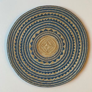 May include: A round, flat woven wall basket featuring a concentric circular pattern. The design alternates between natural tan and deep blue fibers, creating a series of rings and jagged, sawtooth-like geometric shapes radiating from the center.