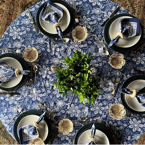 Peut inclure: Une table &agrave; manger ronde dress&eacute;e pour un repas, avec une nappe bleue &agrave; motifs floraux. Chaque couvert comprend une assiette blanche, un dessous de plat vert fonc&eacute;, une serviette bleue &agrave; motifs et un anneau de serviette tiss&eacute;. Des couverts et de petits bols tiss&eacute;s sont &eacute;galement pr&eacute;sents.