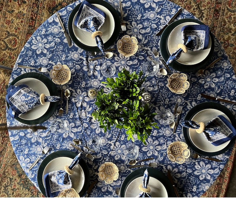 Peut inclure: Une table &agrave; manger ronde dress&eacute;e pour un repas, avec une nappe bleue &agrave; motifs floraux. Chaque couvert comprend une assiette blanche, un dessous de plat vert fonc&eacute;, une serviette bleue &agrave; motifs et un anneau de serviette tiss&eacute;. Des couverts et de petits bols tiss&eacute;s sont &eacute;galement pr&eacute;sents.