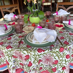 Puede incluir: Un mantel blanco con un patr&oacute;n floral con granadas rosas y hojas verdes. El mantel tiene un borde rosa y azul. Una mesa est&aacute; puesta con servilletas rosas y blancas, vasos transparentes y una maceta verde con orqu&iacute;deas rosas y amarillas.