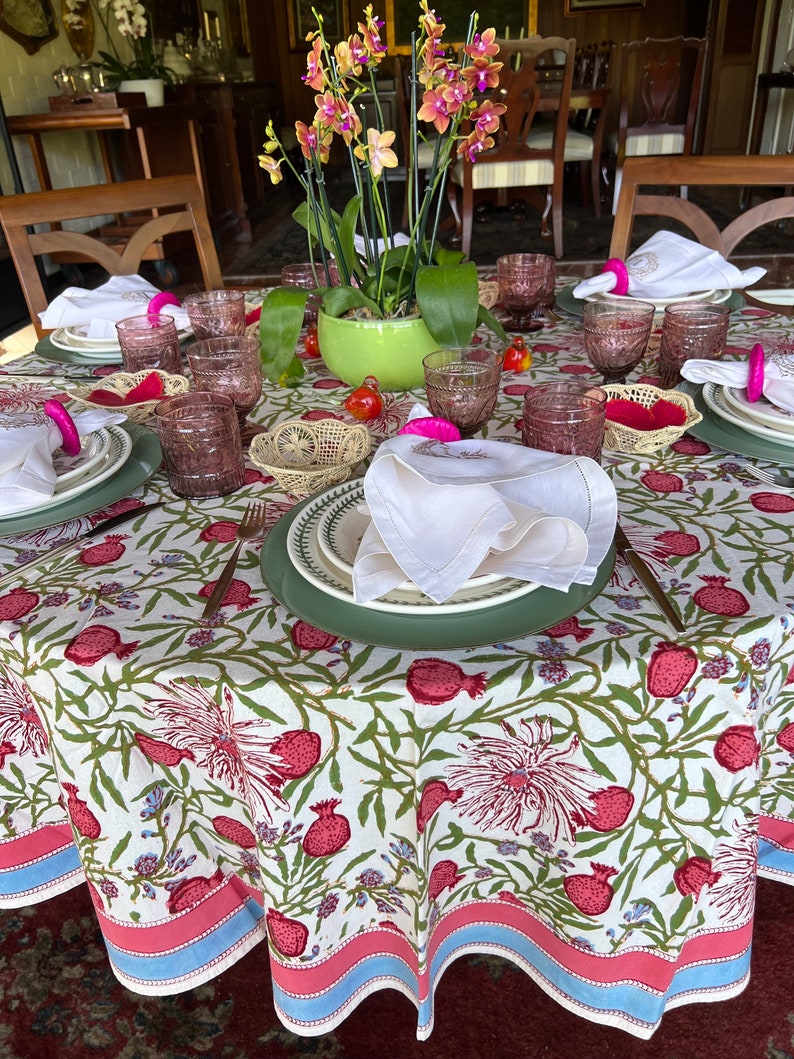 Puede incluir: Un mantel blanco con un patr&oacute;n floral con granadas rosas y hojas verdes. El mantel tiene un borde rosa y azul. Una mesa est&aacute; puesta con servilletas rosas y blancas, vasos transparentes y una maceta verde con orqu&iacute;deas rosas y amarillas.