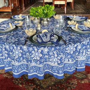 Peut inclure: Une table ronde dress&eacute;e pour un repas, avec une nappe florale bleue et blanche. Chaque couvert comprend une assiette, une serviette et un anneau de serviette tress&eacute;. Des verres transparents et des couverts sont dispos&eacute;s autour de la table, avec un centre de table de verdure.