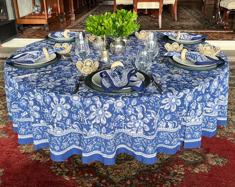 Peut inclure: Une table ronde dress&eacute;e pour un repas, avec une nappe florale bleue et blanche. Chaque couvert comprend une assiette, une serviette et un anneau de serviette tress&eacute;. Des verres transparents et des couverts sont dispos&eacute;s autour de la table, avec un centre de table de verdure.