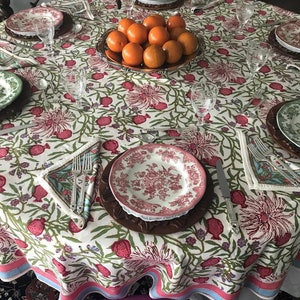 Puede incluir: Un mantel blanco con un patr&oacute;n floral con granadas y hojas verdes. El mantel tiene un borde rosa y azul. Un taz&oacute;n de naranjas se encuentra en el centro de la mesa, rodeado de platos, cubiertos y vasos.