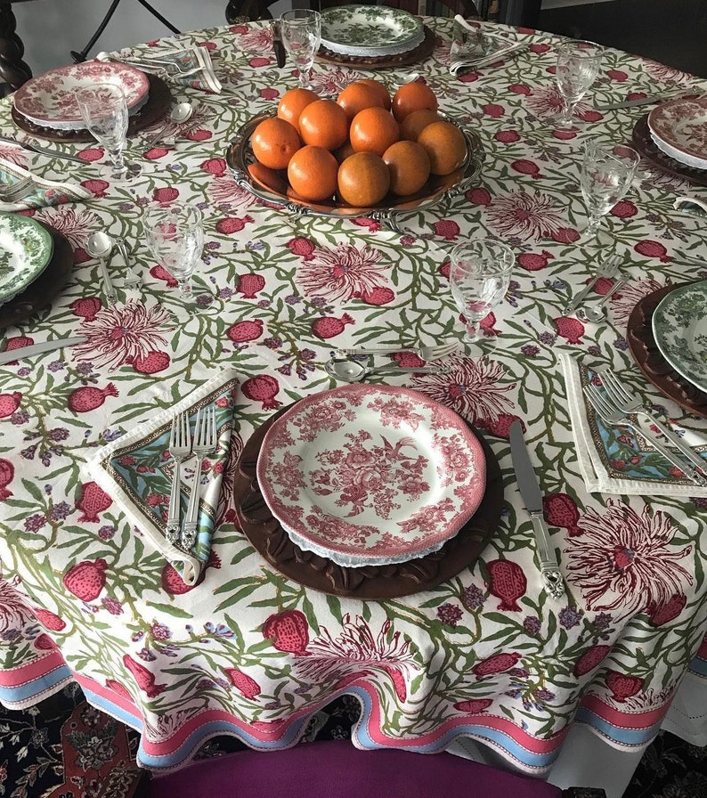 Puede incluir: Un mantel blanco con un patr&oacute;n floral con granadas y hojas verdes. El mantel tiene un borde rosa y azul. Un taz&oacute;n de naranjas se encuentra en el centro de la mesa, rodeado de platos, cubiertos y vasos.