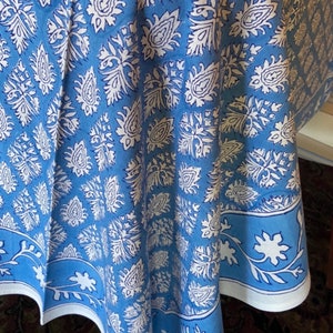 May include: A blue and white patterned tablecloth. The tablecloth features a repeating paisley and floral design in white against a light blue background. The border has a white floral design on a blue background. The fabric is draped over a table.