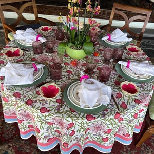 Puede incluir: Un mantel blanco con estampado floral presenta una mesa puesta con servilletas blancas, anillos de servilleta rosas y platos verdes. La mesa est&aacute; decorada con una planta en maceta con flores rosas.