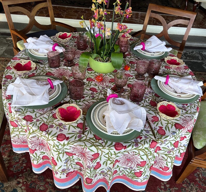 Puede incluir: Un mantel blanco con estampado floral presenta una mesa puesta con servilletas blancas, anillos de servilleta rosas y platos verdes. La mesa est&aacute; decorada con una planta en maceta con flores rosas.