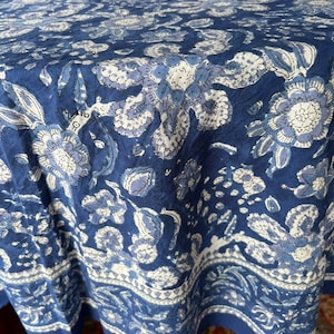 Peut inclure: Une nappe bleue &agrave; motifs floraux avec un motif de fleurs blanches et bleu clair. La nappe a une bordure avec un motif g&eacute;om&eacute;trique r&eacute;p&eacute;titif. Le tissu est drap&eacute;, montrant la texture et les plis du mat&eacute;riau.