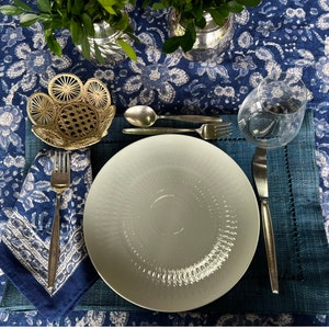 Peut inclure: Une table dress&eacute;e avec une assiette blanche, des couverts et un verre &agrave; eau sur un set de table bleu. La table est recouverte d'une nappe bleue &agrave; motifs floraux. Un petit panier d&eacute;coratif et une serviette pli&eacute;e sont &eacute;galement pr&eacute;sents.