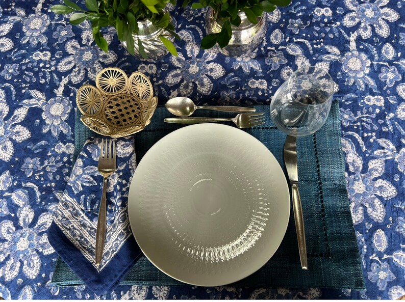 Peut inclure: Une table dress&eacute;e avec une assiette blanche, des couverts et un verre &agrave; eau sur un set de table bleu. La table est recouverte d'une nappe bleue &agrave; motifs floraux. Un petit panier d&eacute;coratif et une serviette pli&eacute;e sont &eacute;galement pr&eacute;sents.
