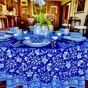 Peut inclure: Une table &agrave; manger ronde, orn&eacute;e d'une nappe bleue vibrante orn&eacute;e d'un motif floral blanc. La table est dress&eacute;e avec des assiettes blanches, des bols bleus &agrave; motifs et des verres transparents. Des couverts dor&eacute;s et un vase d'orchid&eacute;es blanches compl&egrave;tent le cadre &eacute;l&eacute;gant.