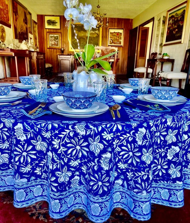 Peut inclure: Une table &agrave; manger ronde, orn&eacute;e d'une nappe bleue vibrante orn&eacute;e d'un motif floral blanc. La table est dress&eacute;e avec des assiettes blanches, des bols bleus &agrave; motifs et des verres transparents. Des couverts dor&eacute;s et un vase d'orchid&eacute;es blanches compl&egrave;tent le cadre &eacute;l&eacute;gant.