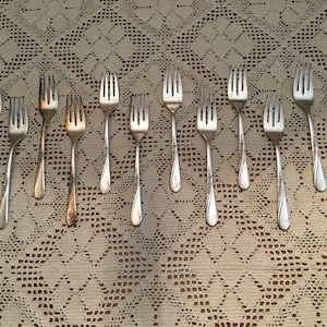 May include: A set of twelve silver forks laid out in two rows on a white lace tablecloth.