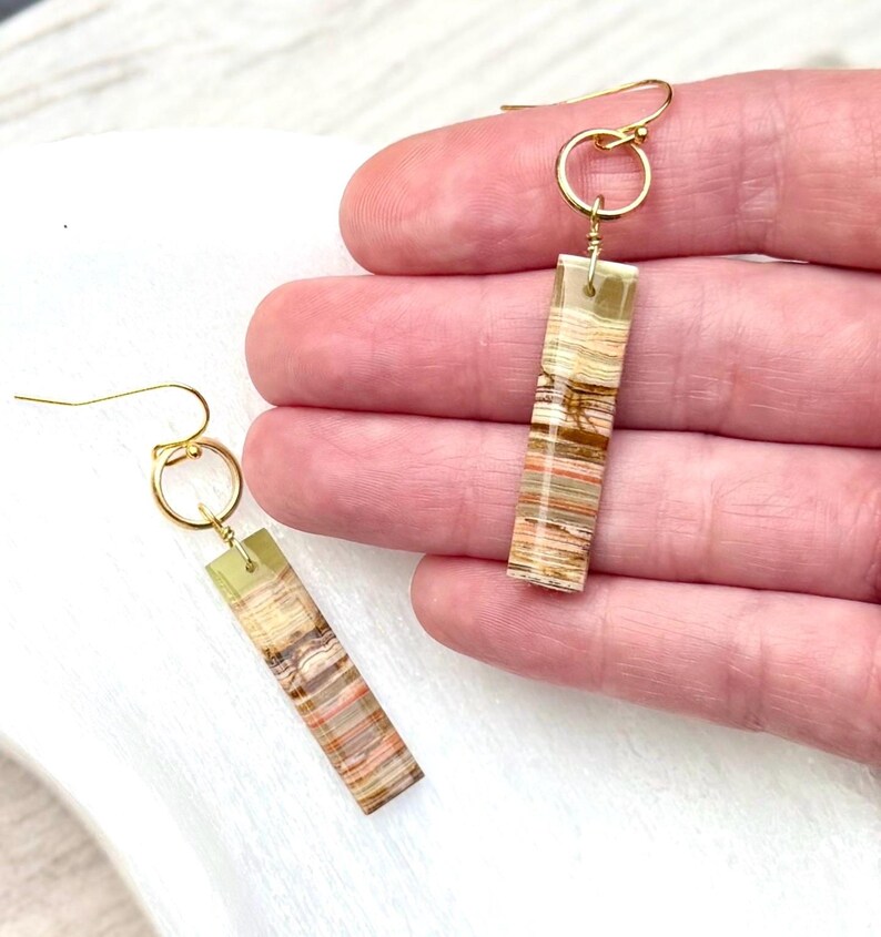 Op de afbeelding: Een paar rechthoekige oorhangers. Elke oorbel heeft een goudkleurige haak en een rechthoekige hanger met lagen in beige, bruin en perziktinten. De oorbellen zijn ontworpen voor doorboorde oren.