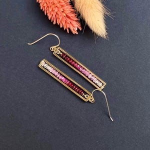 May include: A pair of gold-colored earrings with a rectangular design. Each earring features a row of small, colorful gemstones in shades of pink and red. The earrings have a hook closure and are displayed on a dark blue surface.