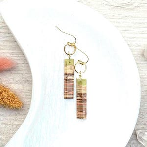 Op de afbeelding: Een paar goudkleurige oorbellen met rechthoekige, gelaagde, aardetinten hangers. De oorbellen hebben een goudkleurige haak en een kleine gouden ring die de haak met de hanger verbindt. De hangers hebben tinten bruin, beige en een vleugje roze.