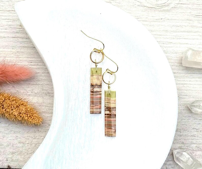 Op de afbeelding: Een paar goudkleurige oorbellen met rechthoekige, gelaagde, aardetinten hangers. De oorbellen hebben een goudkleurige haak en een kleine gouden ring die de haak met de hanger verbindt. De hangers hebben tinten bruin, beige en een vleugje roze.
