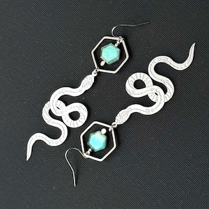 May include: A pair of silver-toned earrings featuring a snake design. Each earring has a hexagonal frame with a turquoise-colored stone and a dangling snake silhouette. The earrings are set against a black background.