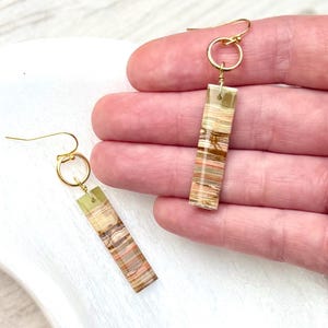 Op de afbeelding: Een paar goudkleurige oorhangers. Elke oorbel heeft een rechthoekige, gelaagde steen in tinten bruin, beige en groen, die is opgehangen aan een goudkleurige ring en oorhaak. De oorbellen worden gepresenteerd tegen een neutrale achtergrond.