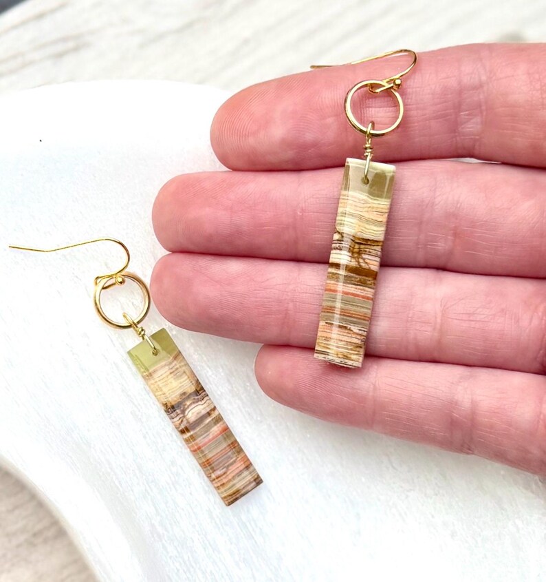 Op de afbeelding: Een paar goudkleurige oorhangers. Elke oorbel heeft een rechthoekige, gelaagde steen in tinten bruin, beige en groen, die is opgehangen aan een goudkleurige ring en oorhaak. De oorbellen worden gepresenteerd tegen een neutrale achtergrond.