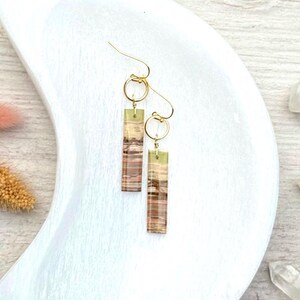 Op de afbeelding: Een paar goudkleurige oorhangers. Elke oorbel heeft een rechthoekige, gestreepte hanger in bruin-, beige- en groentinten, die aan een gouden ring en haak is bevestigd. De oorbellen worden gepresenteerd op een wit, halvemaanvormig oppervlak.