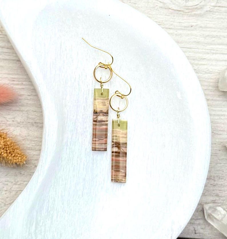 Op de afbeelding: Een paar goudkleurige oorhangers. Elke oorbel heeft een rechthoekige, gestreepte hanger in bruin-, beige- en groentinten, die aan een gouden ring en haak is bevestigd. De oorbellen worden gepresenteerd op een wit, halvemaanvormig oppervlak.