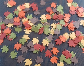 May include: A scattering of autumn-themed paper leaves in shades of brown, orange, yellow, and green. The leaves are in the shape of maple leaves and are scattered on a dark wood surface.