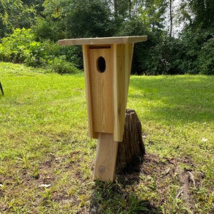 May include: A wooden birdhouse with a slanted roof and a small opening for birds to enter. The birdhouse is mounted on a tree stump in a grassy area.