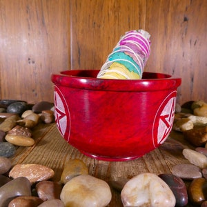 May include: A red, round, stone bowl with a white pentagram design on each side. A white sage smudge stick with colourful ribbons is resting inside the bowl. The bowl is surrounded by smooth, grey, brown, and white stones.