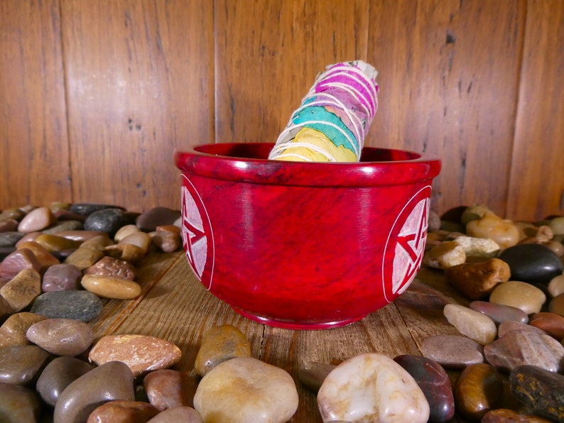 May include: A red, round, stone bowl with a white pentagram design on each side. A white sage smudge stick with colourful ribbons is resting inside the bowl. The bowl is surrounded by smooth, grey, brown, and white stones.