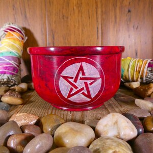 May include: A red, round, stone bowl with a white pentagram carved into the centre. The bowl is surrounded by small, smooth stones.