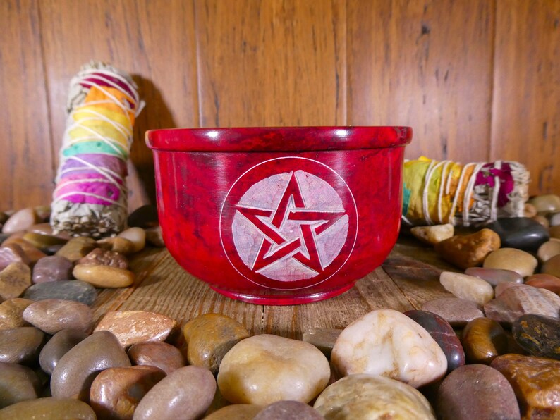 May include: A red, round, stone bowl with a white pentagram carved into the centre. The bowl is surrounded by small, smooth stones.