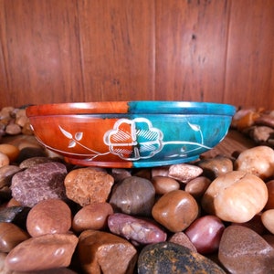 May include: A round, decorative bowl made of carved stone. The bowl is split in half, with one side painted a deep red and the other a teal blue. Both sides feature a white floral design.