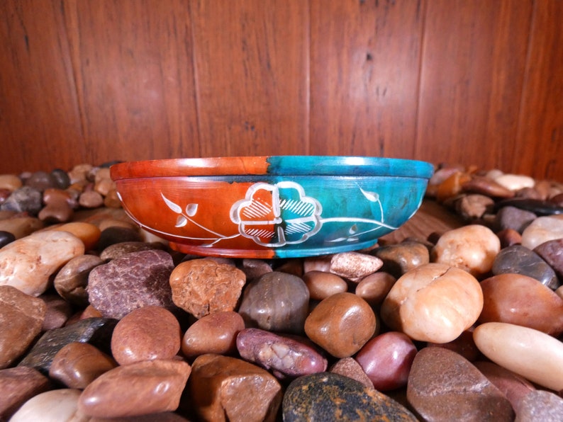 May include: A round, decorative bowl made of carved stone. The bowl is split in half, with one side painted a deep red and the other a teal blue. Both sides feature a white floral design.
