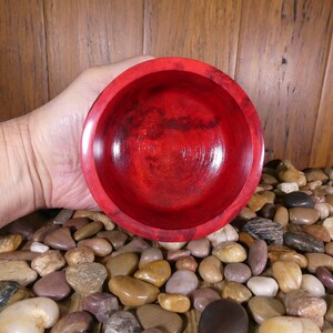 May include: A deep red wooden bowl with a smooth, polished finish. The bowl is held in a hand, with a background of small, smooth stones.