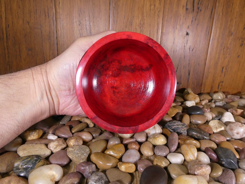May include: A deep red wooden bowl with a smooth, polished finish. The bowl is held in a hand, with a background of small, smooth stones.