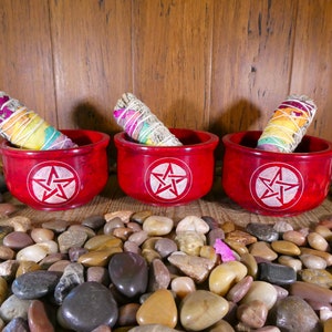 May include: Three red, round, wooden bowls with a white pentagram design. Each bowl contains a smudge stick with white sage and colourful flower petals.