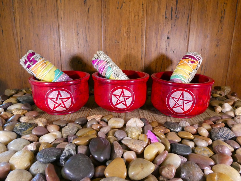May include: Three red, round, wooden bowls with a white pentagram design. Each bowl contains a smudge stick with white sage and colourful flower petals.