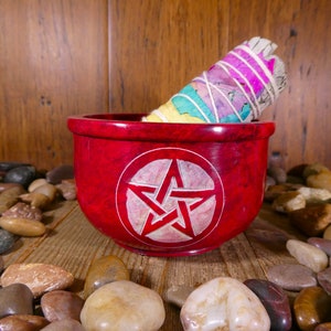 May include: A red, round, stone bowl with a white pentagram carved into the centre. A white sage smudge stick is resting on the rim of the bowl. The bowl is surrounded by smooth, grey and brown stones.